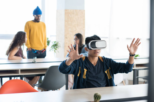 Content handsome young Asian male app developer in casual clothing sitting at table and gesturing hands while using virtual reality simulator in coworking space - Photo, Image
