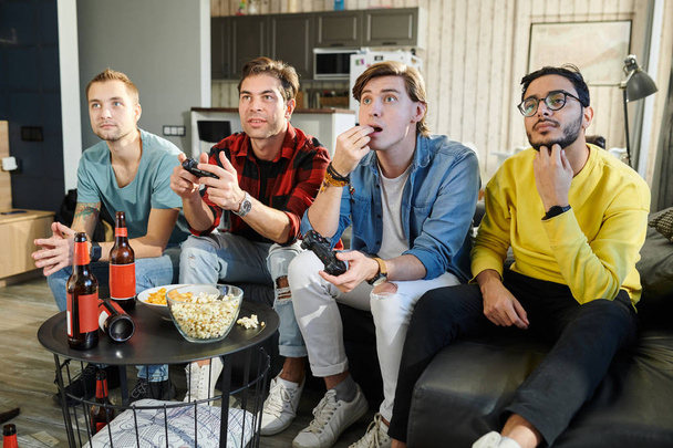 Group of young men sitting on sofa eating popcorn and drinking beer and looking forward to the results of the game - Photo, Image