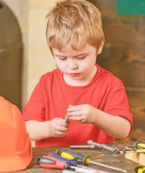 Closeup kid playing with metal bolts. Little laborer in workshop. Cute boy in red T-shirt behind the table - Fotó, kép