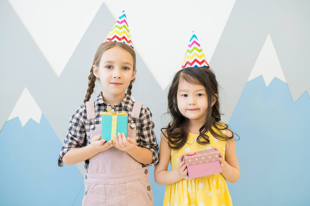 Content cute multi-ethnic little girls in colorful party hats standing against painted wall and holding small gift boxes while looking at camera - Photo, Image