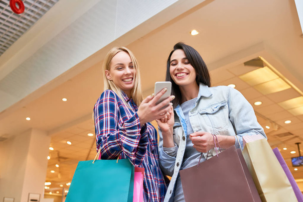 Gelukkig jonge vrouwen staan met boodschappentassen en het lezen van een bericht samen op mobiele telefoon in het winkelcentrum - Foto, afbeelding