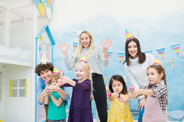 joyeuses mères excitées en vêtements décontractés s'amusant avec les enfants à la fête d'anniversaire : enfants heureux éclatant craquelins dans la salle de jeux décorées guirlandes drapeau
 - Photo, image
