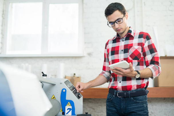 Випадковий молодий чоловік в окулярах дивиться планшет і використовує сучасне поліграфічне обладнання в кабінеті типографії
 - Фото, зображення