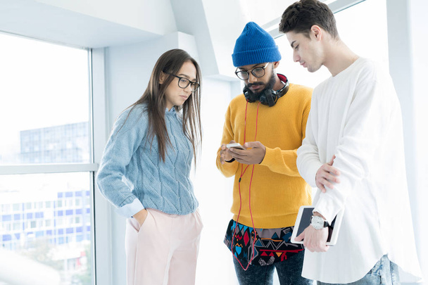 Studenti hipster seri in abiti casual in piedi in corridoio e guardare video su smartphone mentre discutono del progetto
 - Foto, immagini