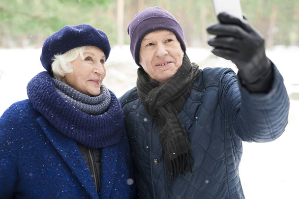 Portrait of modern senior couple taking selfie photo while enjoying walk in winter park - Photo, Image