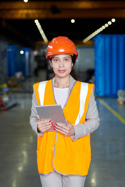 Porträt einer Fabrikarbeiterin mit Hut und reflektierender Weste, die in die Kamera blickt und fröhlich lächelt, während sie in der Werkstatt des Produktionsbetriebs posiert, Kopierraum - Foto, Bild