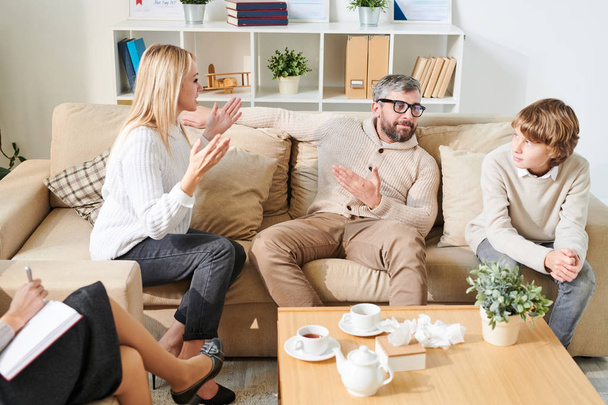 Serious handsome bearded father in glasses pointing at emotional mother in hysteria while explaining reason of divorce to teenage son at therapy session - Photo, Image