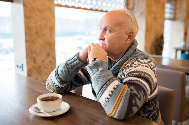 Σοβαρός γεροντικός ηλικιωμένος άνθρωπος με ζεστό πουλόβερ που κάθεται σε ξύλινο τραπέζι με κούπα καφέ και γέρνει το κεφάλι στα χέρια, ενώ κοιτάζοντας έξω παράθυρο στο μοντέρνο καφέ - Φωτογραφία, εικόνα