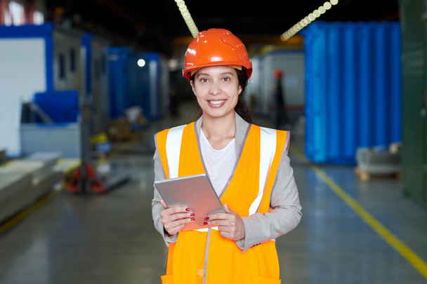 Taillenhoch Porträt einer Fabrikarbeiterin mit Hut und reflektierender Weste, die in die Kamera blickt und glücklich lächelt, während sie in der Werkstatt der Produktionsanlage posiert, Kopierraum - Foto, Bild