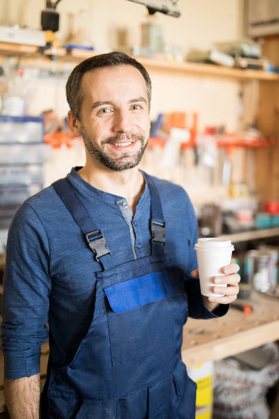 Retrato de la cintura hacia arriba del carpintero maduro mirando a la cámara y sonriendo mientras toma un descanso de café en el trabajo
 - Foto, Imagen