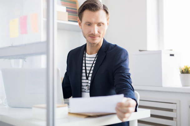 Porträt eines erwachsenen Geschäftsmannes, der Dokument liest, während er am Schreibtisch im Büro sitzt, Kopierraum - Foto, Bild