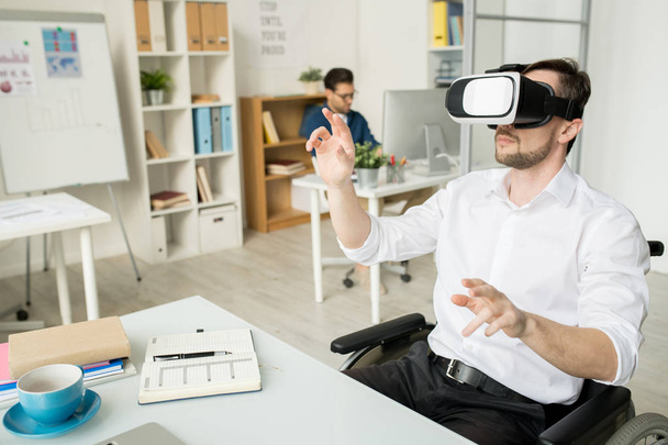 Young man in formalwear sitting in wheelchair wearing virtual reality headset and gesturing while his colleague working in the background - Foto, imagen