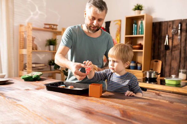 Content handsome father and cute little son standing at kitchen counter and putting heart-shaped cookie into box while they making gift for mother - Zdjęcie, obraz