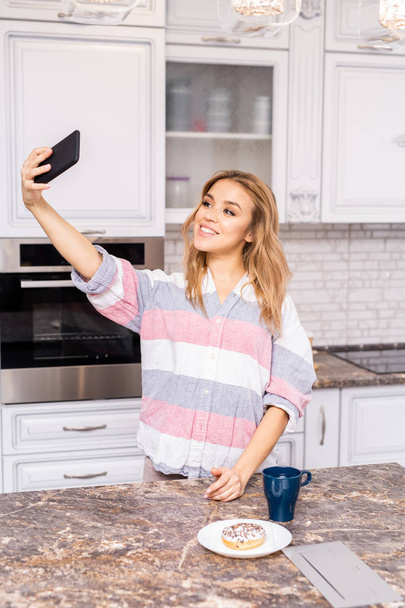 Cintura até retrato de bela jovem mulher tomando selfie na cozinha enquanto desfruta de café da manhã saudável na cozinha
 - Foto, Imagem
