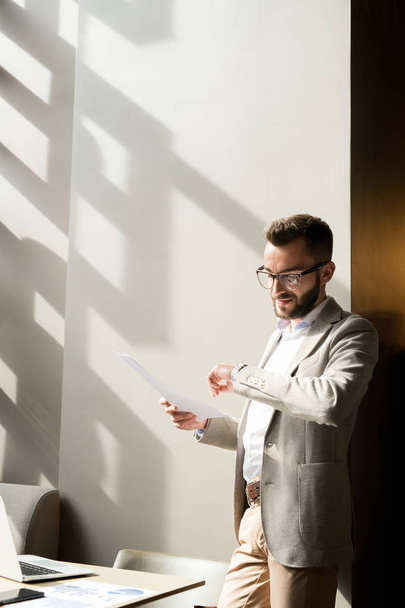 Jonge zelfverzekerde zakenman staat op kantoor met document en kijkend naar zijn horloge, hij wacht op een ontmoeting - Foto, afbeelding
