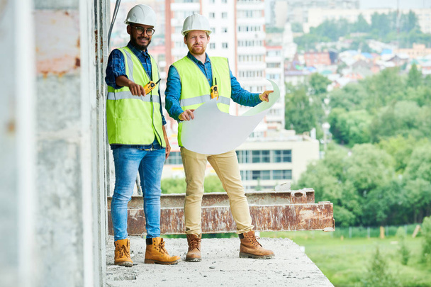 Due uomini in berretti rigidi e gilet che tengono il progetto mentre ispezionano l'edificio incompiuto in cantiere
 - Foto, immagini
