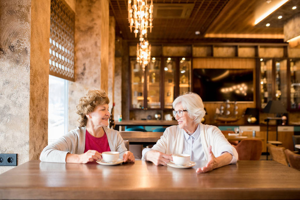Positivo atraente seniores conversadores senhoras em cardigans sentado à mesa de madeira no café e discutir últimas notícias enquanto bebem café juntos
 - Foto, Imagem