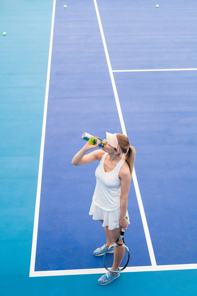 High angle portrait of young blonde woman drinking water from sports bottle in indoor tennis court, copy space - Photo, Image