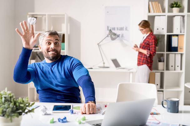 positiver verspielter gutaussehender bärtiger Programmierer im Pullover, der am Tisch sitzt und Papierkugeln wirft, während er Zeit im modernen Büro verschwendet - Foto, Bild