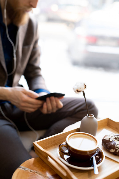 Close-up of wooden tray with morning cappuccino and chocolate doughnut, hipster man using smartphone in background - Photo, Image
