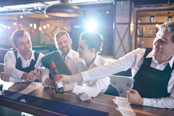 Ομάδα των θετικών ευτυχείς σερβιτόρους σε ομοιόμορφη που γέρνει σε πάγκο μπαρ και κουδουνίζουν μπουκάλια μπύρας, ενώ Γιορτάζοντας την επιτυχία μετά από ώρες στο εστιατόριο - Φωτογραφία, εικόνα