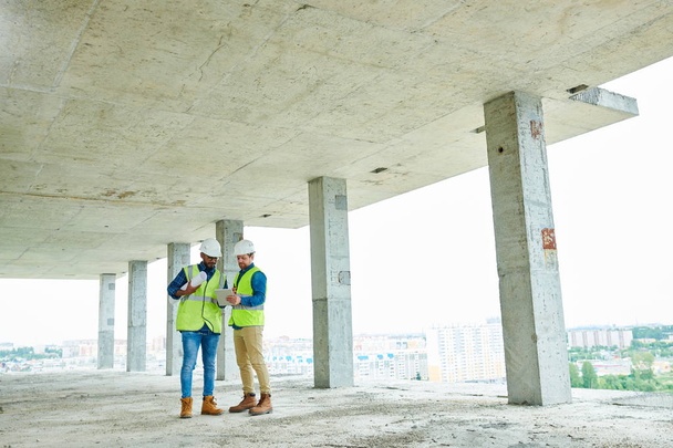 Két férfi védősisakok és mellények megvitatása építési terv mellett állva felső határa befejezetlen épület - Fotó, kép