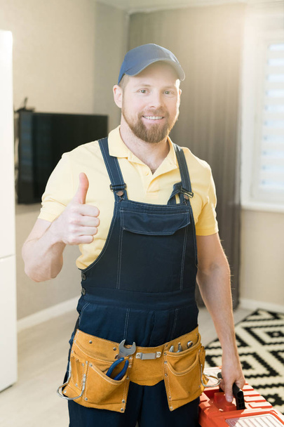 Veselý, vzrušený mladý vousatý údržbář v modrém čepici a džínovu celkově ukazující na nářadí a na přidržující skříňku s nářadím při pohledu na fotoaparát v moderním bytě. - Fotografie, Obrázek