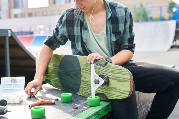Mid-section portrait of contemporary young man fixing skateboard changing wheels on bench at skate park - Photo, image