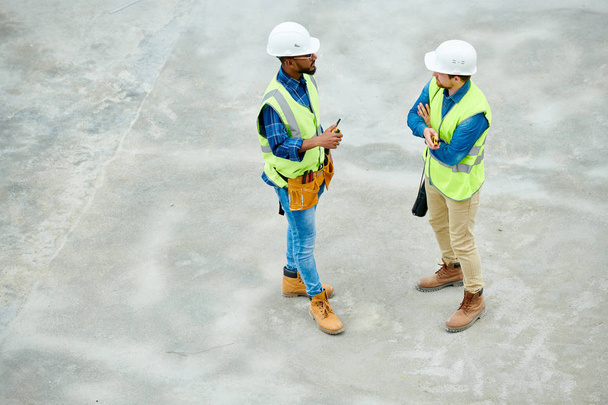Side view of two men in hardhats and waistcoats standing on construction site and talking with each other during inspection - Photo, Image