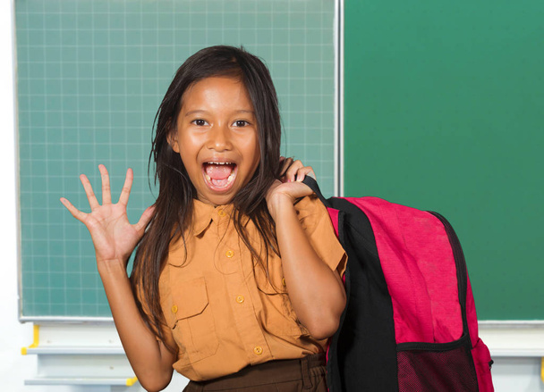 beautiful happy and excited female child in school uniform carrying student bag smiling cheerful standing at classroom blackboard in back to school and kid education - Foto, immagini