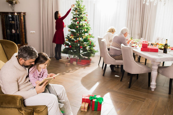 Escena de familia disfrutando juntos del día de Navidad, madre decorando el árbol mientras su marido lee cuentos a los niños
 - Foto, imagen