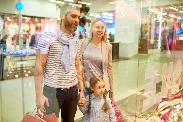 Portrait of happy family looking at window displays while shopping in mall together, shot from behind glass - Foto, imagen