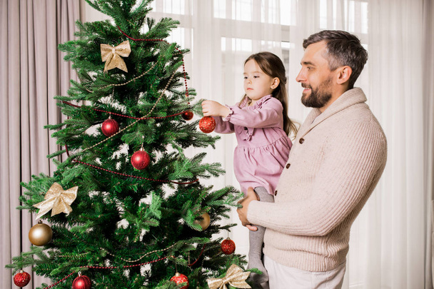 Baba ile ev dekorasyon sırasında Noel ağacı üzerinde süsler asılı sevimli küçük kız Yan görünüm portre, kopya alanı - Fotoğraf, Görsel