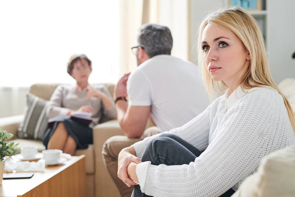 Sad thoughtful attractive young wife with blond hair sitting on sofa and embracing her knees while her husband talking to psychiatrist at therapy session - Photo, Image