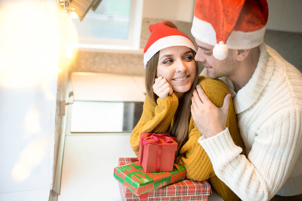 Homem abraçando mulher bonita ambos vestindo chapéus de Papai Noel de pé na cozinha com presentes
 - Foto, Imagem