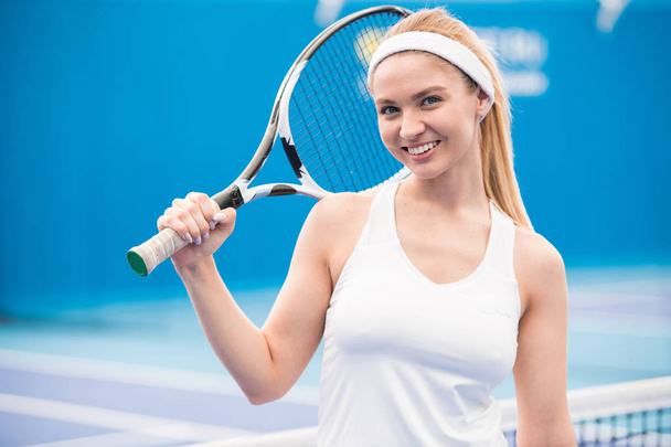 Waist up portrait of cheerful female tennis player holding racket posing  in indoor court and looking at camera smiling happily, copy space - Foto, imagen
