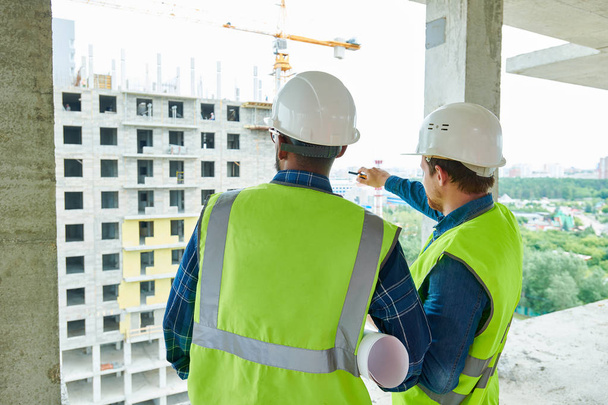 Задний вид строителей в касках и зеленых жилетах, обсуждающих структуру здания, мужчина-инженер, указывающий на строящееся здание
 - Фото, изображение