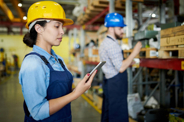 Retrato de vista lateral de una trabajadora de fábrica que usa un sombrero de señora usando una tableta digital en el taller con otro trabajador en segundo plano, espacio para copiar
 - Foto, Imagen