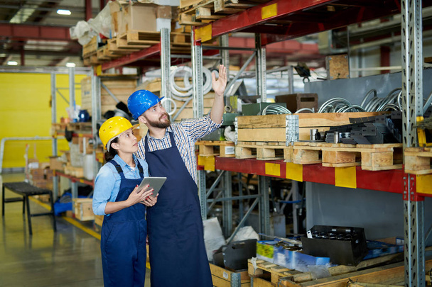 Portrait of two modern factory workers, man and woman, wearing hardhats doing inventory standing by tall shelves in warehouse pointing up, copy space - Photo, Image