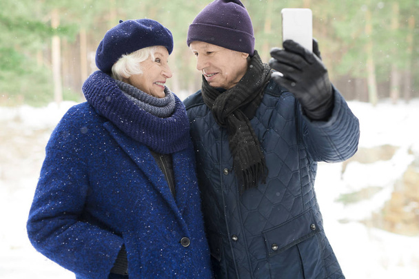 Kış parkında yürüyüş yaparken selfie fotoğrafı çeken sevgi dolu yaşlı çiftin bel denyukarı portresi - Fotoğraf, Görsel
