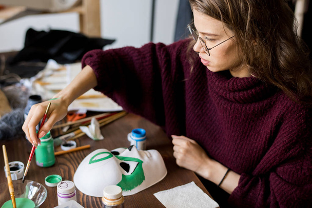 Nahaufnahme einer ernsten, konzentrierten jungen Frau in Brille, die am Schreibtisch sitzt und im Designstudio eine Maske mit grüner Farbe malte, Halloween-Kostüm - Foto, Bild