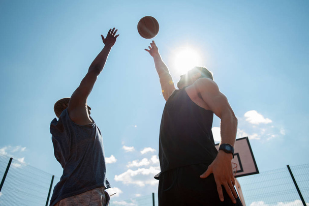 Χαμηλή γωνία θέα δύο ανδρών που παίζουν μπάσκετ και άλμα φθάνοντας μπάλα εναντίον του γαλάζιου ουρανού, αντιγραφή χώρου - Φωτογραφία, εικόνα