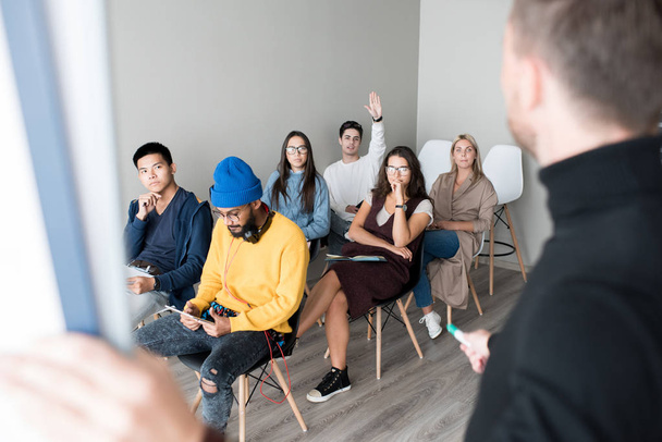 Young audience at training class: handsome man raising hand to ask question, other students sitting on chairs and talking to speaker in convention center - Photo, Image