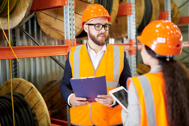 Portrait of modern bearded businessman wearing hardhat listening to factory employee while standing in workshop and holding clipboard, copy space - Foto, immagini