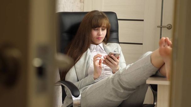 beautiful businesswoman with phone sits in a chair with bare feet on the table resting. girl at work in the office keeps a note on a mobile phone. - Photo, Image