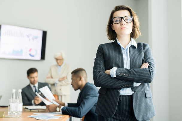 Serious purposeful attractive young female business executive in formal suit crossing arms on chest and looking at camera while business team discussing papers in background - Photo, Image