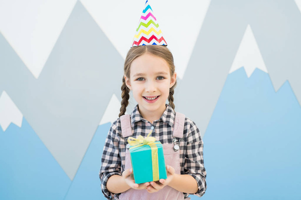 Jolly excited little girl in zigzag-lined party hat standing against creative wall with mountains drawings and holding gift box while looking at camera - Foto, afbeelding