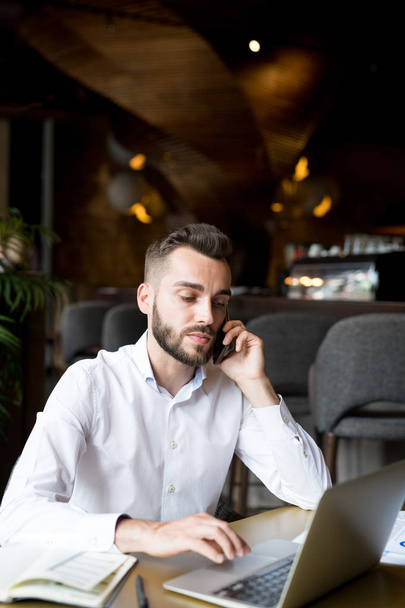 Retrato de un joven hombre de negocios barbudo hablando por teléfono mientras trabajaba con una computadora portátil en la cafetería
 - Foto, Imagen