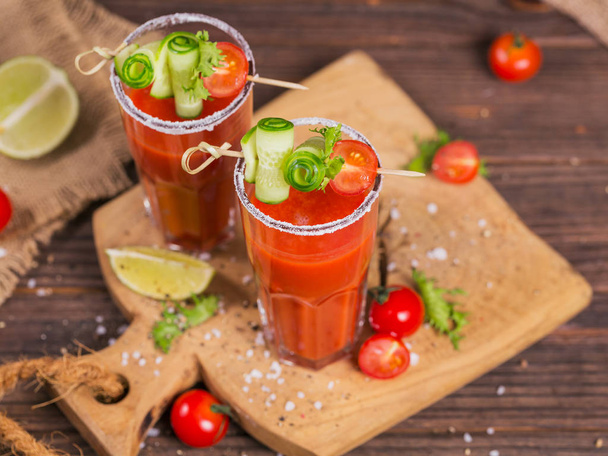 Two glasses of fresh organic tomato juice decorated with raw tomatoes, cucumber and leaves on a rustic wooden cutting board - Фото, изображение