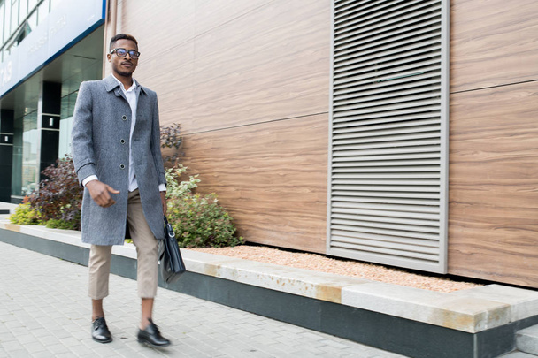 Σοβαρός όμορφος μοντέρνος νέος Αφρικανός επιχειρηματίας φορώντας γκρι παλτό και κουβαλάει χαρτοφύλακα ενώ περπατάει με σιγουριά στην οικονομική περιοχή, φεύγει από το γραφείο - Φωτογραφία, εικόνα